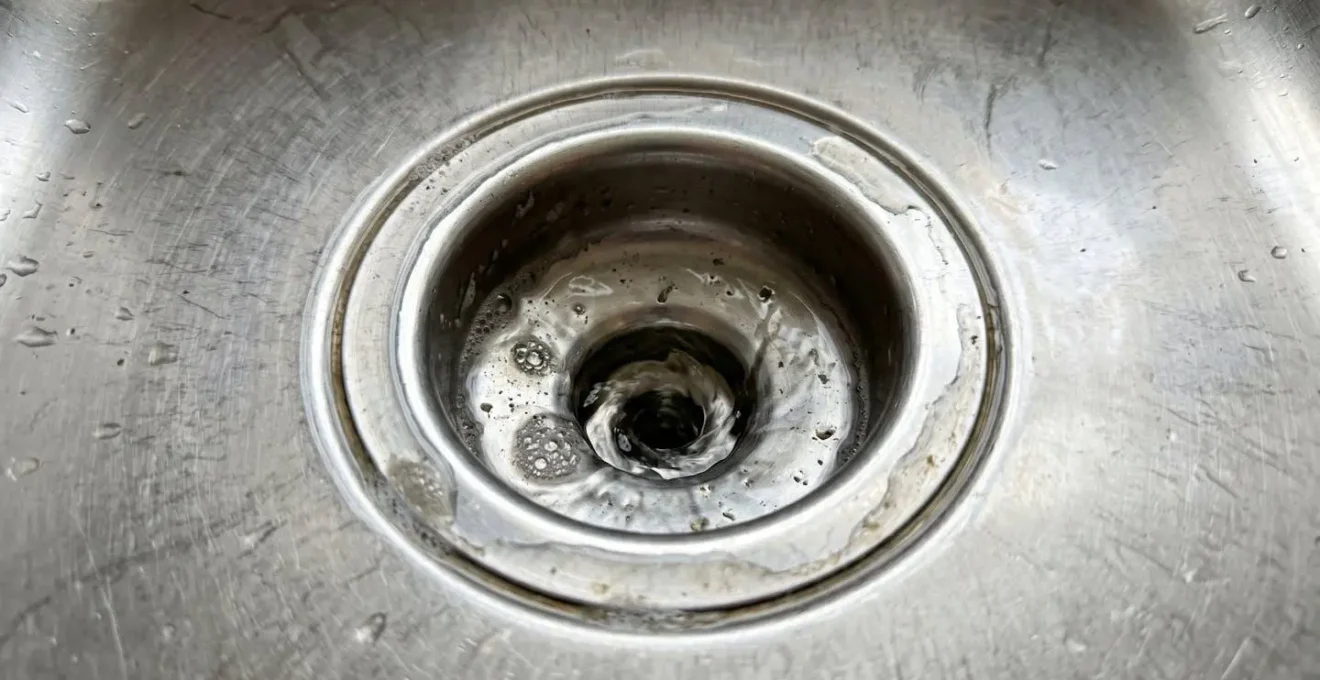 Close-up view of a kitchen sink drain with water pooling slowly around the opening, showing visible drainage delay